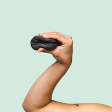 Aperson's hand holding a small, dark, textured, konjac sponge against a solid pale mint green background.