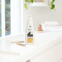 Ecostore Ultra Power 3x Concentrated Laundry Liquid with Wooden Pegs on a Bench and Brush and Folded Laundry in the Background. Soft Lighting from a Window and a Plant in the Background.